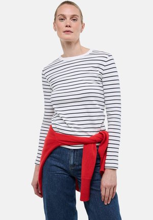 Femme blonde portant une chemise blanche à manches longues avec des rayures horizontales noires, un jean bleu et un pull rouge noué autour de la taille.