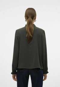 Donkergroene blouse met lange mouwen van textuurstof, met een gerimpelde kraag en knoopmanchetten. Gecombineerd met donkerdenim broek.