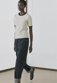 Frau läuft drinnen, trägt ein weißes und schwarzes, gestreiftes Kurzarmshirt, dunkle, umgeschlagene Jeans und schwarze Slipper.