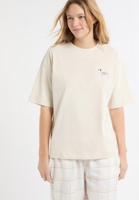 Beżowa koszulka z krótkim rękawem wykonana z miękkiej tkaniny, z małym haftowanym wzorem psa na piersi. Do tego jasne spodnie w kratę.