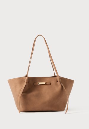 Bruine leren totebag met twee lange hengsels, taps toelopende zijkanten en een vlakke bodem. Voorzien van een horizontale metalen accenten aan de voorkant.