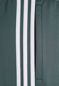 Pantaloni da track in tessuto verde scuro con tre strisce bianche lungo il lato. Include una piccola tasca a sinistra. Materiale testurizzato.