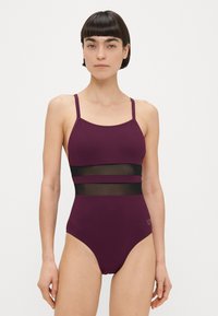 Γυναίκα με κοντά σκούρα μαλλιά που φοράει μονοκόμματο μαγιό σκούρου μοβ χρώματος, με δύο οριζόντια διάφανα μαύρα πλέγματα στη μέση.