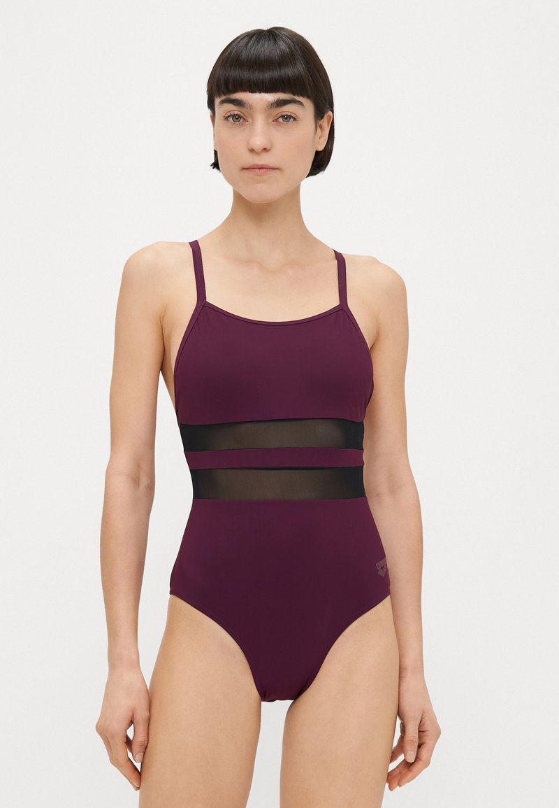 Γυναίκα με κοντά σκούρα μαλλιά που φοράει μονοκόμματο μαγιό σκούρου μοβ χρώματος, με δύο οριζόντια διάφανα μαύρα πλέγματα στη μέση.