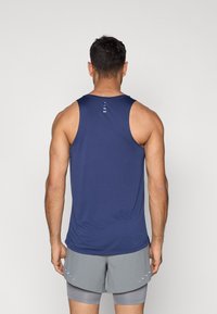 Maiou sport albastru marin cu o textură netedă, având o detaliere în spate cu trei dungi albe. Combinat cu șorturi gri.