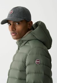 Huzatos olajzöld dzseki kapucnival, steppelt textúrával és kerek hímzett logóval. Szürke gyapjúsapka teszi teljessé a megjelenést.