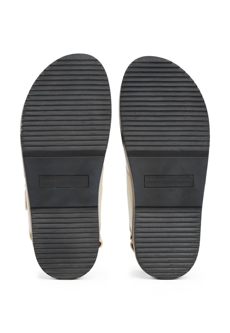 Schwarze Gummisohlen mit vertikalen Rillen, beiges Obermaterial, mit einem eingeprägten Logo im Fersenbereich. Einfaches, flaches Design.