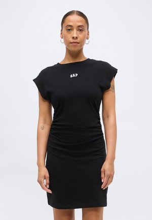 Žena s kruhovými náušnicami, oblečená v čiernych šatách bez rukávov s logom "GAP" na hrudi, stojaca pred jednoduchým svetlým pozadím.