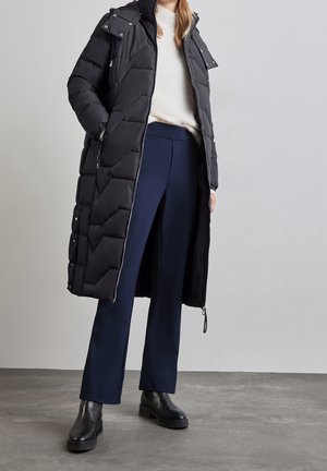 Frau trägt langen schwarzen Steppmantel mit Kapuze, cremefarbenen Pullover, marineblaue Hose und schwarze Stiefeletten, steht auf grauem Boden.