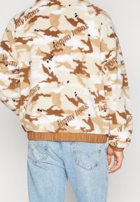 Giacca in pile con motivo camouflage beige e marrone, caratterizzata da orlo elastico a coste, colletto alto e sottili accenti di branding.