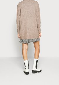 Beige stickad kofta, lång och oversize, bärs över en svart och vit mönstrad, volangklänning. Ett par vita ankelskor med svarta detaljer.