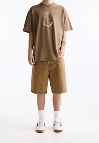Camiseta de algodón marrón con un diseño de cara sonriente y texto, combinada con pantalones cortos de mezclilla color beige y zapatillas blancas con acentos en marrón.