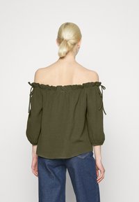 Blusa verde-oliva com decote à off-the-shoulder, gola com babados, mangas com laço e tecido texturizado. Usada com calças de ganga azuis.