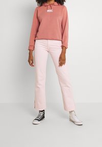 Felpa color rosa polvere con cordoncino e logo stampato, abbinata a jeans flared rosa chiaro con fessure laterali, indossata con sneakers in bianco e nero.