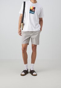 Uomo che indossa una t-shirt bianca con grafica, pantaloni corti a strisce beige e neri, calzini bianchi, sandali marroni e una borsa a tracolla beige, in piedi di fronte a un muro semplice.