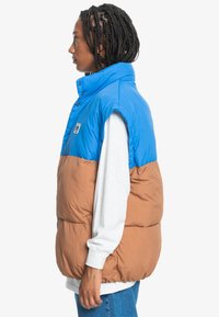 Colorful puffer vest featuring a blue upper section and brown lower section, with a high collar and visible stitching. Snap button closure.