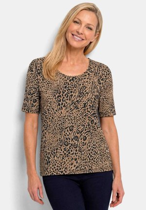 Frau lächelt, trägt ein kurzärmeliges Oberteil mit Leopardenmuster und dunkle Hose, steht vor einem schlichten hellen Hintergrund.