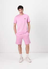 Rosa bomulls t-shirt med korta ärmar och en liten gul fjärilslogotyp på bröstet, matchad med rosa shorts. Vita sneakers.