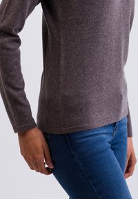 Brun stickad tröja med långa ärmar och rundad hals, kombinerad med blå skinny jeans. Texturen verkar mjuk och något strukturerad.