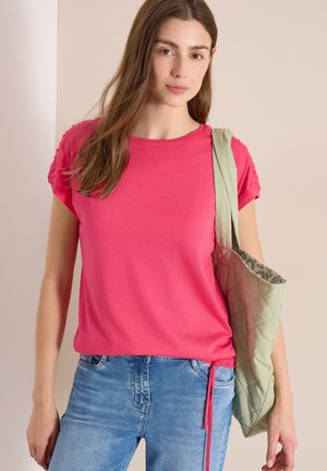 Jeune femme portant un haut à manches courtes rose vif et un jean bleu clair, avec un sac à bandoulière matelassé vert clair.