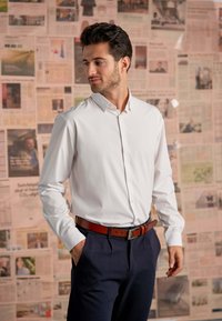 Hvid skjorte med lange ærmer og krave med knap, kombineret med navy bukser og et brunt læderbælte. Baggrunden har sider fra aviser.