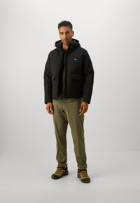 Homme portant une doudoune noire, un pantalon vert olive et des chaussures de randonnée beiges, se tenant contre un fond gris clair uni.