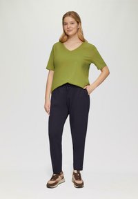 Vrouw met blond haar in een groene V-hals korte mouw top, zwarte broek en bruine sneakers, staande met één hand in haar zak.
