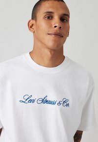 Weißes Baumwoll-T-Shirt mit blauer Stickschrift "Levi Strauss & Co." auf der Vorderseite. Kurze Ärmel und runder Ausschnitt.