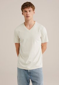 V-puoleinen, lyhythihainen t-paita vaaleasta beige puuvillasta, jossa on rento leikkaus ja sileä pinta, yhdistettynä vaaleansinsisiin farkkuihin.