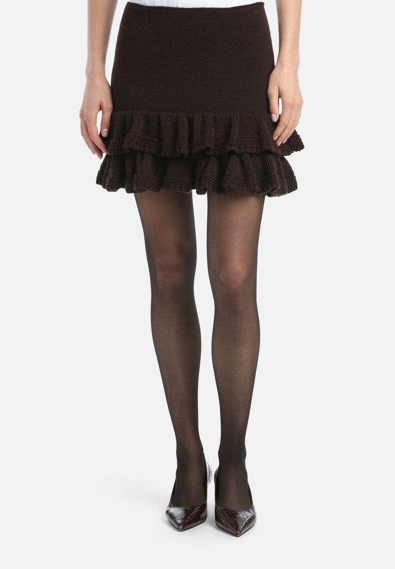 Burgunderfarbener Strickrock mit schichtigem, rüschengesäumtem Saum. Die Trägerin trägt transparente schwarze Strumpfhosen und spitz zulaufende, metallische High Heels.