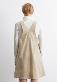 Beige kordflod klänning med hängslen som korsar över ryggen, en lös passform och en slät textur. Bärs över en långärmad vit topp.