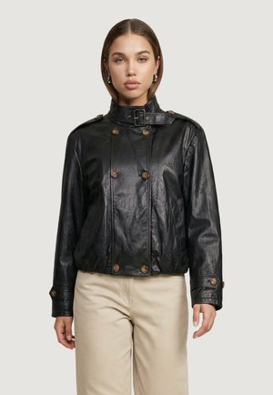 Donna che indossa una giacca nera in eco-pelle con bottoni e colletto con fibbia e pantaloni beige, in piedi su uno sfondo uniforme.