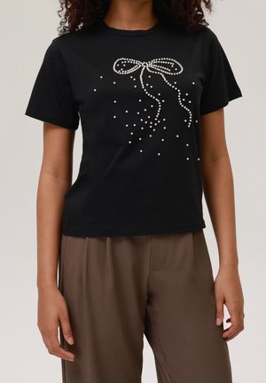 Kvinde iført sort t-shirt med hvid perlebue-design og spredte perler, kombineret med brune bukser med høj talje mod ensfarvet baggrund.