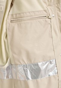 Beżowa odzież wierzchnia z miękką polarem, z odblaskową srebrną taśmą i kieszenią na zamek. Tekst zawiera symbole pielęgnacji i znakowanie.