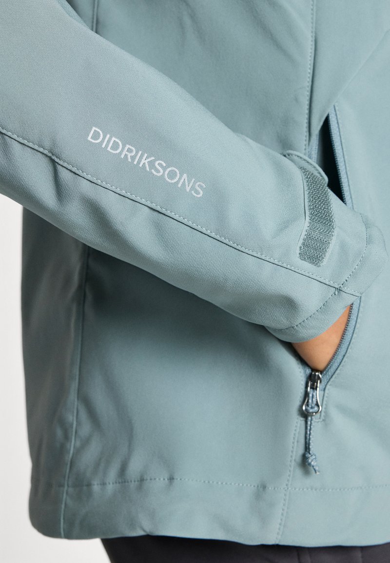 Himmelblauer Didriksons-Jackenärmel mit Markenname, Hand in der seitlichen Zipp-Tasche und verstellbarem Handgelenkriemen mit Klettverschluss-Detail.
