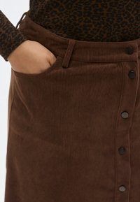 Fustă maro din catifea, cu nasturi în față, având un buzunar lateral și o textură striată, purtată cu un top cu model.