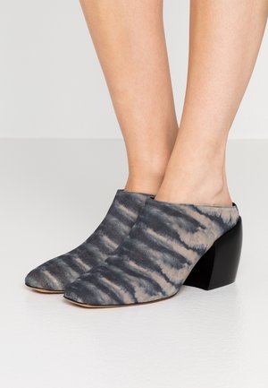 Stopy w noszonych na nogach butach mule na koturnie z szaroczarną tkanką w stylu tie-dye i masywnymi czarnymi obcasami, stojące na białej podłodze.