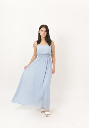 Jeune femme aux cheveux raides et foncés portant une robe longue sans manches bleu clair et des chaussures blanches ouvertes à l'avant, debout devant un fond uni.