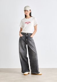 Jeans de pierna ancha grises combinados con una camiseta crema que presenta el logo rojo "TOMMY JEANS". Una gorra de color claro completa el atuendo.