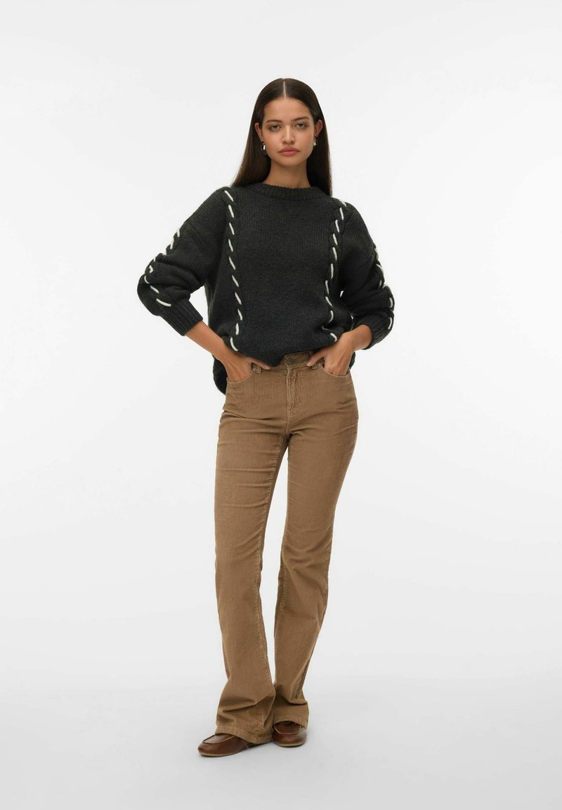 Maglione grigio scuro con accenti di cucitura bianca, abbinato a pantaloni in corduroy beige a zampa d'elefante. La modella si trova in piedi con le mani sui fianchi su uno sfondo bianco.