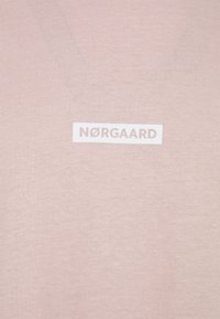 Světle růžové tričko vyrobené z jemné tkaniny, s bílým obdélníkovým logem s nápisem "NØRGAARD" tučně napsaným přes střed.