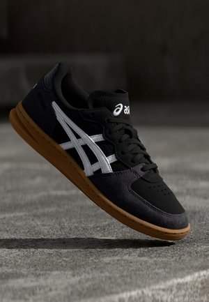 Sort-hvid Asics-sneaker med gummisål hænger over en betongulv i svagt lys.