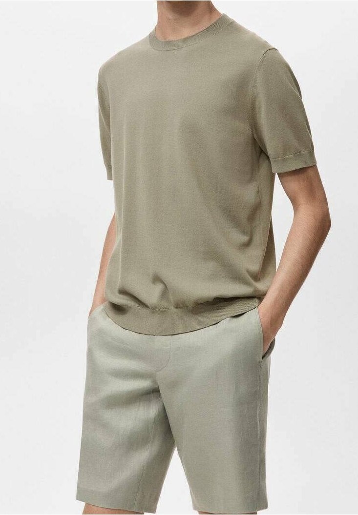 Homme portant un t-shirt en maille à manches courtes olive clair et un short assorti olive clair longueur genou, les mains dans les poches devant un fond uni.