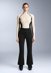 Krémový žebrovaný top s vysokým límcem a černými bočními panely, spárovaný s černými širokými kalhotami s páskem v pase a rozparkem vpředu.