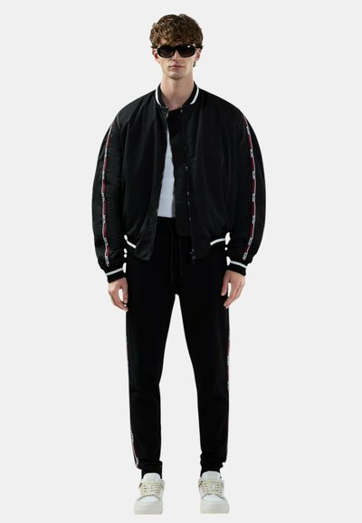 Młody mężczyzna ubrany w czarną kurtkę typu bomber oraz dopasowane spodnie dresowe, białe sneakersy i ciemne okulary przeciwsłoneczne, stojący na białym tle.