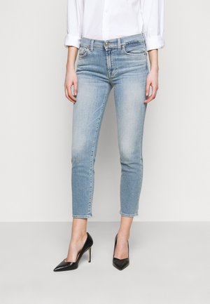 Persoon draagt lichtblauwe cropped skinny jeans, wit overhemd met lange mouwen met opgerolde manchetten, en zwarte puntige hoge hakken.