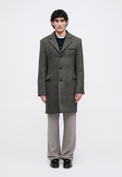Homme portant un long manteau en chevrons gris, une chemise bleu marine, un pantalon gris clair et des chaussures noires, se tenant devant un fond clair uniforme.