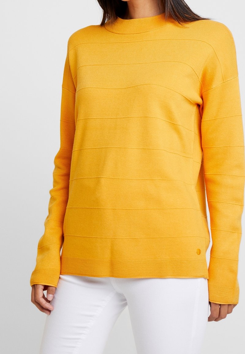 Femme portant un pull moutarde à manches longues avec de subtils rayures horizontales texturées et un pantalon blanc, sur un fond uni.