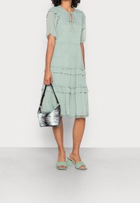 Frau trägt ein hellgrünes Midi-Kleid mit Punkten und Rüschen, hält eine Handtasche mit schwarz-weißem Muster und trägt passende grüne High Heels.
