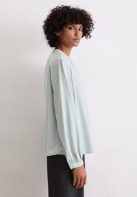 Jasnoniebieska, bluzka z długim rękawem o luźnym kroju, z guzikami przy mankietach i okrągłym dekoltem. Miękka tkanina o minimalnej fakturze.